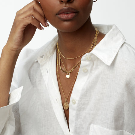 model in a white shirt wearing a gold bead chain choker necklace, a gold padlock charm on gold figaro necklace, a gold snake chain necklace and a gold athena coin charm on a gold bead chain necklace
